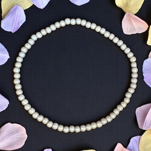 Könnte beinhalten: Eine Halskette aus kleinen, runden, cremefarbenen Perlen, die einen Kreis bilden. Die Halskette befindet sich vor einem schwarzen Hintergrund, mit verstreuten pastellfarbenen Blütenblättern in Lavendel, Rosa und Gelb.