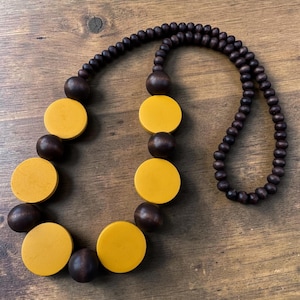 May include: A necklace featuring a series of dark brown spherical wooden beads interspersed with larger, flat, circular mustard yellow wooden discs. The beads are arranged in a continuous strand, alternating between the dark brown spheres and yellow discs.
