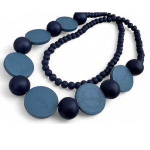 Puede incluir: Un collar azul marino y azul claro con una mezcla de cuentas redondas y en forma de disco. El collar presenta una doble hebra de pequeñas cuentas redondas y cuentas más grandes, de estilo madera, en varios tamaños y formas. El collar está sobre un fondo blanco.
