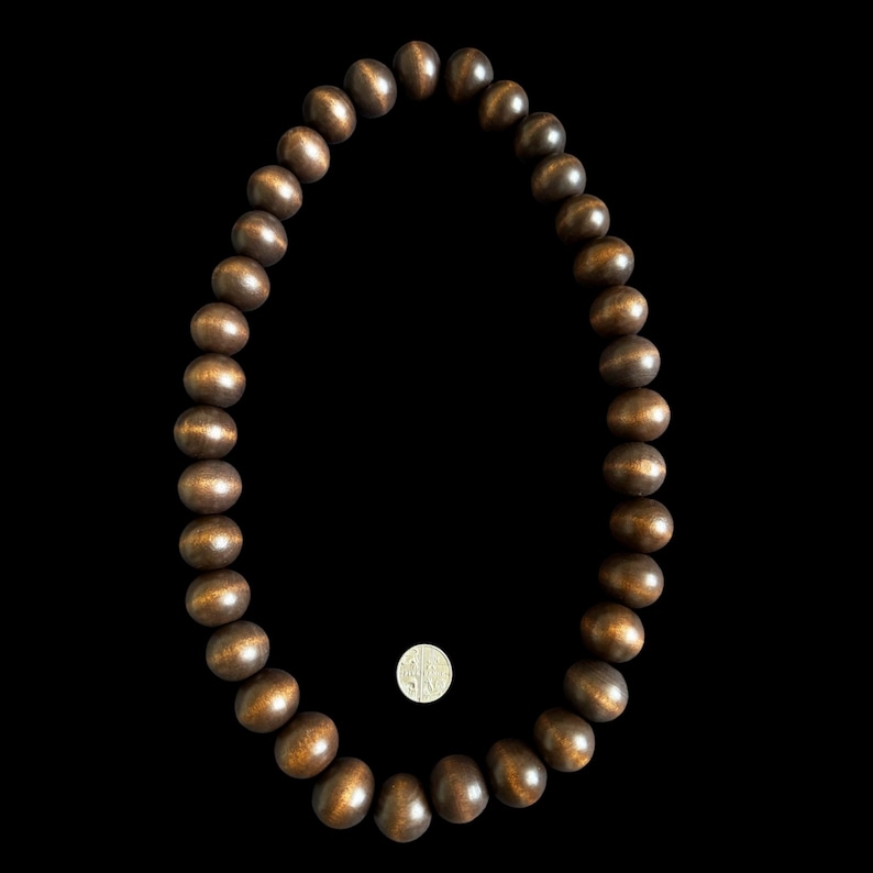 May include: A necklace composed of large, round, dark brown wooden beads. The beads have a polished, glossy finish. A coin is placed near the necklace for size comparison. The necklace is set against a black background.