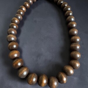 May include: A necklace featuring large, dark brown wooden beads. The beads have a smooth, polished finish and are strung together to form a circular shape. The necklace is displayed against a dark background, highlighting the natural wood grain and color.