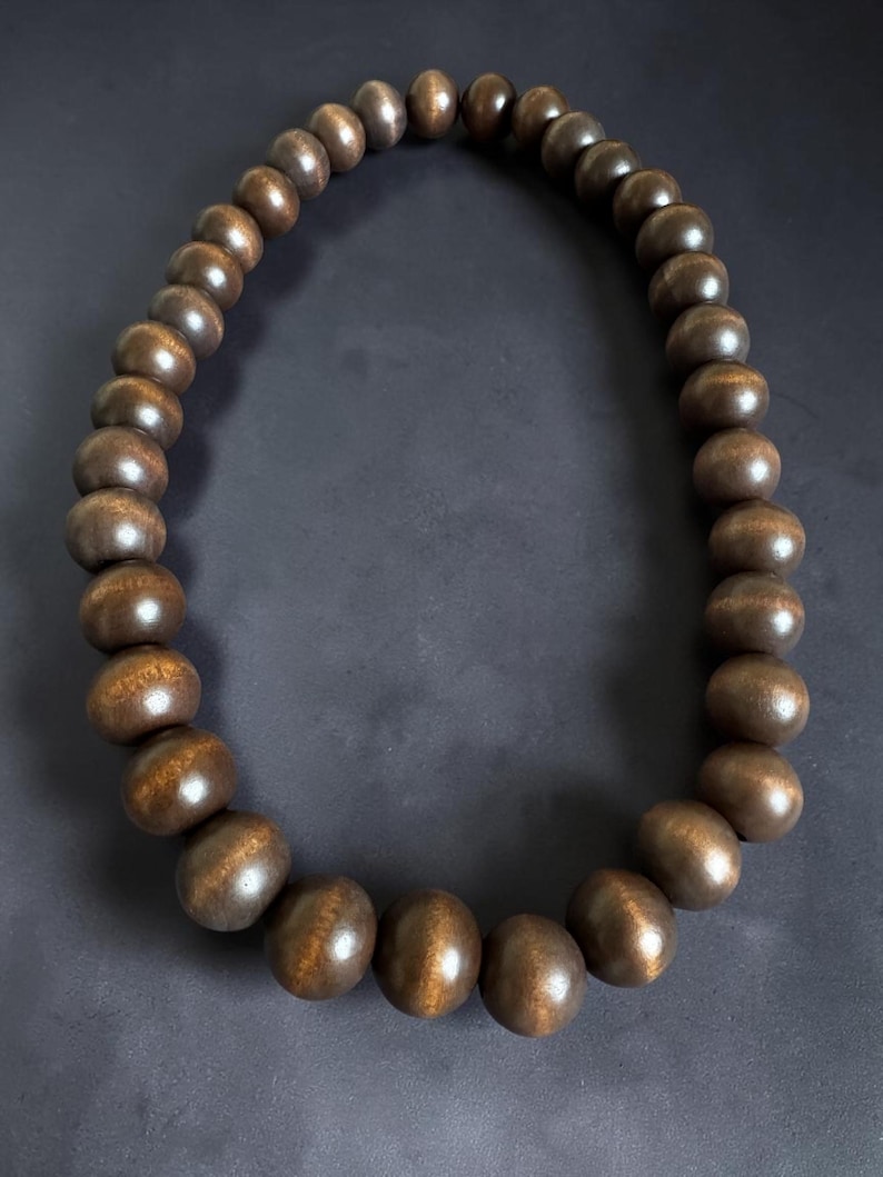 May include: A necklace featuring large, dark brown wooden beads. The beads have a smooth, polished finish and are strung together to form a circular shape. The necklace is displayed against a dark background, highlighting the natural wood grain and color.