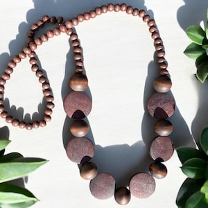 May include: A brown wooden beaded necklace with large, flat, round beads. The necklace is on a white surface with green plants in the background.