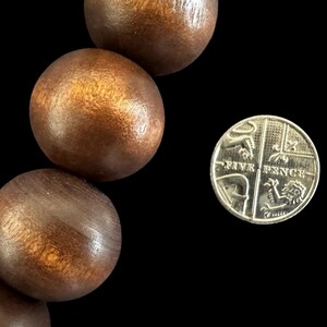 May include: Close-up of large, round, wooden beads with a warm brown finish. A silver Five Pence coin is next to the beads. The beads have a smooth, polished appearance, and the coin shows intricate details.