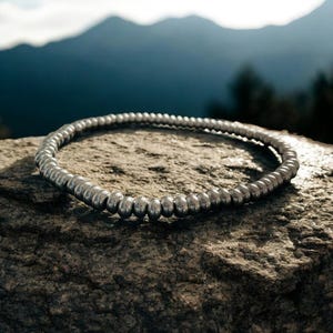 May include: A silver beaded necklace or bracelet, lying on a rough, grey rock surface. The background is a blurry image of mountains and sky.