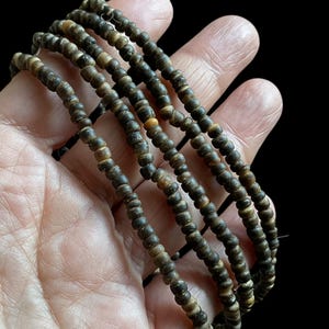 Op de afbeelding: Een streng kleine, ronde, donkerbruine kralen met lichtere bruine en bruingele variaties. De kralen zijn aan elkaar geregen en vormen meerdere strengen. De achtergrond is een donkere, effen kleur.