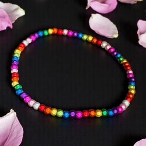 May include: A rainbow-colored beaded bracelet. The beads transition through the colors of the rainbow, from red to purple. The bracelet is on a black surface, with pink flower petals scattered around it.