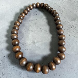 May include: A necklace composed of numerous, evenly-sized, dark brown wooden beads. The beads are strung together to form a circular shape. The wooden beads have a smooth, polished finish, and the necklace is displayed against a gray background.