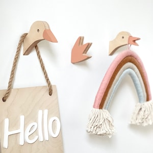 Könnte beinhalten: Wanddekoration aus Holz mit einem Schild, auf dem "Hello" steht, das an einem Holzhaken in Form einer Ente hängt. An einem anderen Holzhaken in Form einer Ente hängt ein Regenbogen-Makramee, und an der Wand befindet sich eine kleine Holzblume.