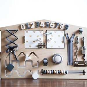 May include: Wooden activity board for toddlers with a house shape, featuring a zipper, a latch, a lock, a chain, a bead maze, a lacing activity, a knob, a bell, and a set of numbers 0-9.