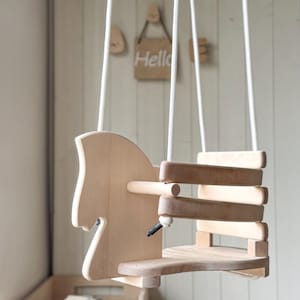 May include: A wooden horse swing with a natural finish. The swing has a seat with a back and a rope hanging from the top.
