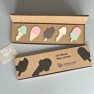 Pode incluir: Um conjunto de cinco ganchos de parede de madeira em forma de cone de sorvete em várias cores, incluindo verde, rosa, marrom, branco e azul claro. Os ganchos são embalados em uma caixa de papelão marrom com o texto "ICE CREAM WALL HOOKS" e "HANDMADE IN LITHUANIA" impresso nela.