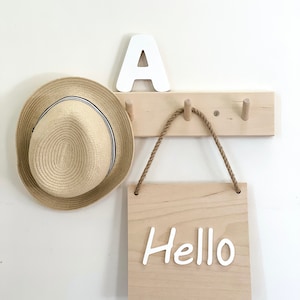 Può includere: Un attaccapanni in legno da parete con tre pioli, un cappello di paglia con una fascia blu navy, una lettera "A" bianca e un cartello di legno con la scritta "Hello" in lettere bianche. Il cartello è appeso a una corda.