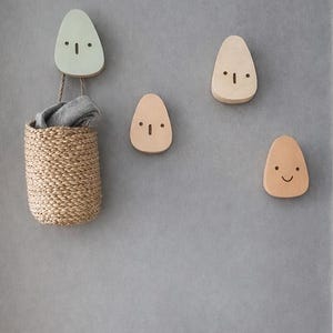 May include: A wall-mounted storage set featuring four wooden face-shaped hooks in varying shades of beige and mint green. A woven basket hangs from one hook, holding a folded gray cloth. A wooden bench sits below the hooks, against a gray wall.