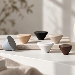 May include: A collection of six conical cabinet knobs in various finishes, including light gray, natural wood, matte black, beige, white, and dark brown. Each knob features a flat circular top and a tapered base, arranged in a group.