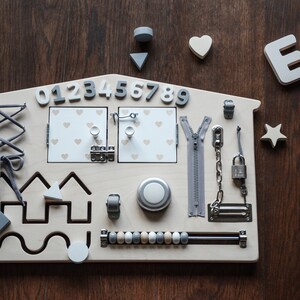 May include: A wooden activity board shaped like a house with various textures and activities for children to explore. It features a zipper, a lock and chain, a latch, a knob, a lacing activity, a bead maze, a shape sorter, and a number sequence from 0 to 9. The board is painted in a light wood color with gray accents.