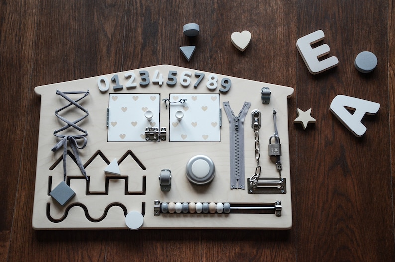 May include: A wooden activity board shaped like a house with various textures and activities for children to explore. It features a zipper, a lock and chain, a latch, a knob, a lacing activity, a bead maze, a shape sorter, and a number sequence from 0 to 9. The board is painted in a light wood color with gray accents.