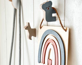 May include: Three wooden wall hooks shaped like a llama, a kangaroo, and a koala. The koala hook supports a square wooden plaque featuring a layered rainbow design in shades of gray, white, and terracotta, with a small heart at the center.