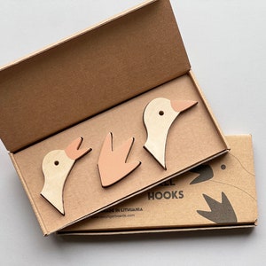 Op de afbeelding: Drie houten gansvormige haken in een kartonnen doos. De haken zijn in beige en roze tinten. De doos heeft de tekst "Goose Hooks" erop gedrukt.