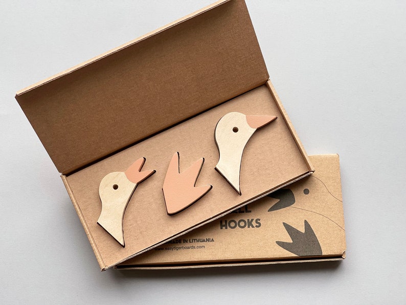 Op de afbeelding: Drie houten gansvormige haken in een kartonnen doos. De haken zijn in beige en roze tinten. De doos heeft de tekst "Goose Hooks" erop gedrukt.