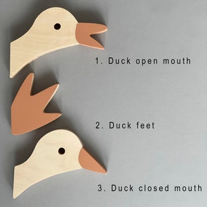 Op de afbeelding: Drie houten eendenkopstukken, geschilderd in tinten bruin en beige. Het eerste stuk heeft een open snavel, het tweede stuk zijn alleen de eendenpoten en het derde stuk heeft een gesloten snavel. De stukken zijn gelabeld met 1. Eend open snavel, 2. Eendenpoten en 3. Eend gesloten snavel.