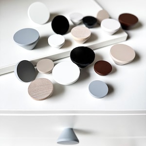 May include: A collection of various wooden cabinet knobs in different colors and shapes. The knobs are in shades of white, black, gray, brown, and natural wood. Some are round, while others have a conical shape. The knobs are arranged on a white surface.