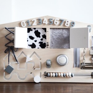 May include: A wooden activity board shaped like a house with various textures and activities for children. It features a zipper, a latch, a lock, a knob, a sliding door, a cow print fabric flap, a gray fabric flap, a bead maze, a shape sorter, and a set of numbers 0-9.