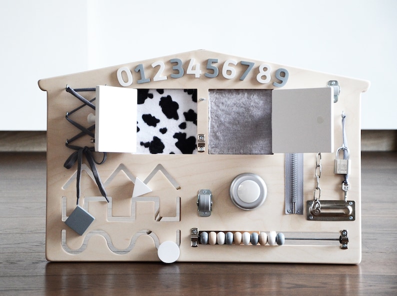 May include: A wooden activity board shaped like a house with various textures and activities for children. It features a zipper, a latch, a lock, a knob, a sliding door, a cow print fabric flap, a gray fabric flap, a bead maze, a shape sorter, and a set of numbers 0-9.