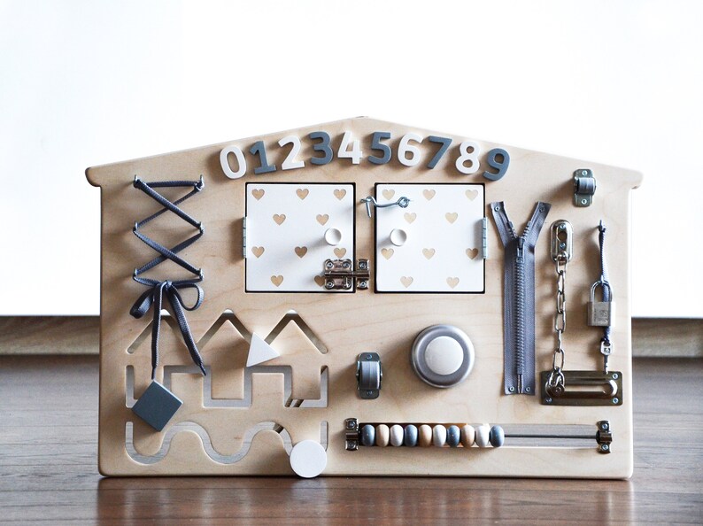 May include: Wooden activity board for toddlers with a house shape, featuring a zipper, a latch, a lock, a chain, a bead maze, a lacing activity, a knob, a bell, and a set of numbers 0-9.