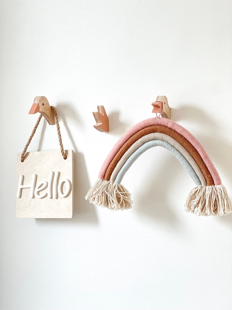 Op de afbeelding: Een houten regenboog wanddecoratie met een lichtroze, lichtbruine en lichtblauwe kleurstelling. De regenboog is gemaakt van garen en heeft aan beide uiteinden een franje. De regenboog hangt aan twee houten vogelhaken. Een houten bord met het woord "Hello" hangt aan een houten vogelhaak.