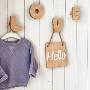 Puede incluir: Un suéter de manga larga en tejido de punto gofrado color morado claro cuelga de un gancho de pared de madera en forma de L. Arriba hay ganchos de madera con forma de letras O, V y E. Un letrero cuadrado de madera con el texto blanco Hello cuelga.