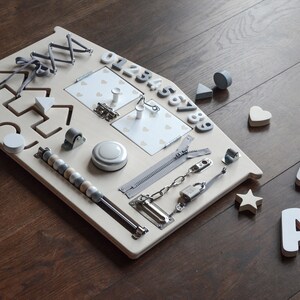 May include: A wooden activity board with various textures and activities for children. It features a zipper, a latch, a chain, a roller, a bell, a bead maze, a shape sorter, a number puzzle, and a mirror. The board is painted in a light beige color and has a gray trim.