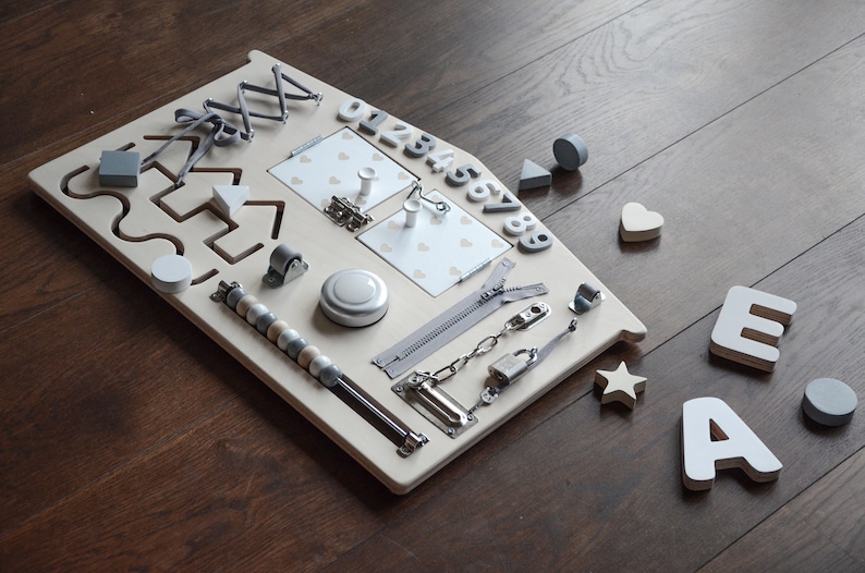 May include: A wooden activity board with various textures and activities for children. It features a zipper, a latch, a chain, a roller, a bell, a bead maze, a shape sorter, a number puzzle, and a mirror. The board is painted in a light beige color and has a gray trim.