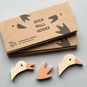 Op de afbeelding: Een set van drie houten eenden wandhaken. De haken zijn geverfd in een lichte houtkleur met roze accenten. De verpakking is van bruin karton met de tekst "Duck Wall Hooks" en "Handmade in Lithuania".