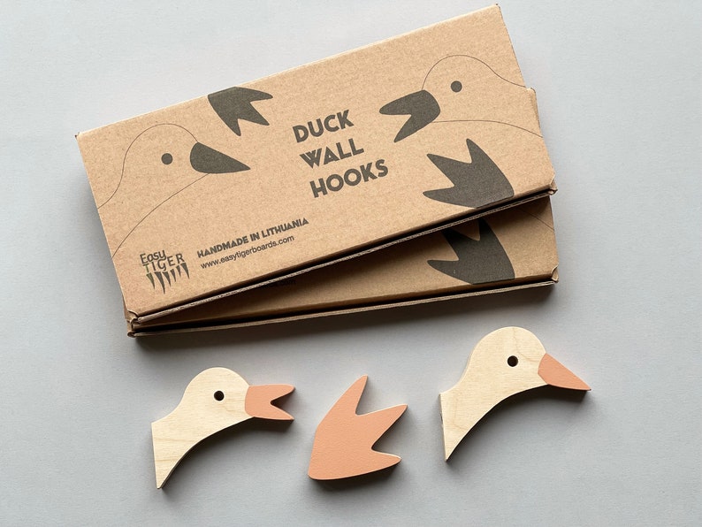 Op de afbeelding: Een set van drie houten eenden wandhaken. De haken zijn geverfd in een lichte houtkleur met roze accenten. De verpakking is van bruin karton met de tekst "Duck Wall Hooks" en "Handmade in Lithuania".