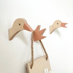 Op de afbeelding: Twee houten gansvormige wandhaken met een roze snavel en een houten bord met de tekst "Hello" die aan de haken hangt met een touw.