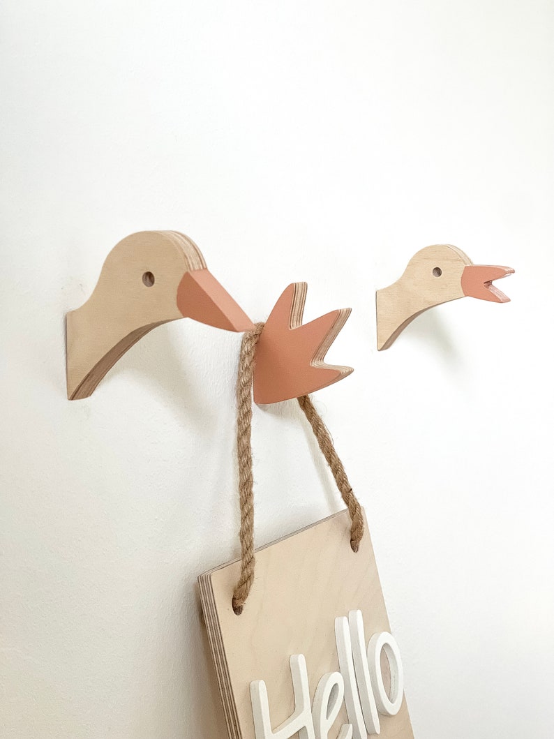 Op de afbeelding: Twee houten gansvormige wandhaken met een roze snavel en een houten bord met de tekst "Hello" die aan de haken hangt met een touw.