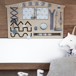 May include: A wooden activity board for children with a house shape, featuring a zipper, a latch, a lock, a bead maze, a lacing activity, and a set of numbers 0-9. The board is mounted on a wall and has a white background.
