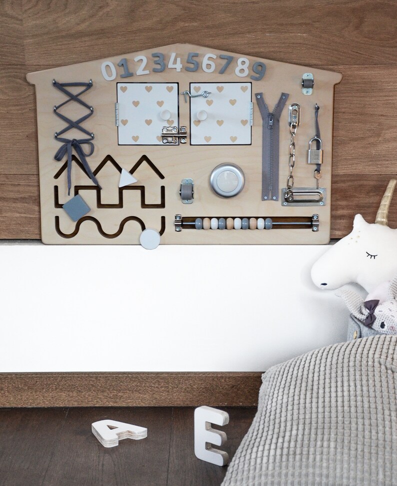 May include: A wooden activity board for children with a house shape, featuring a zipper, a latch, a lock, a bead maze, a lacing activity, and a set of numbers 0-9. The board is mounted on a wall and has a white background.