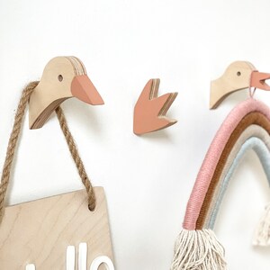 Op de afbeelding: Drie houten wandhaken in de vorm van ganzen en een hand, met een houten bord waarop "Hello" staat, dat aan een van de haken hangt. Een regenboogkleurige garenwandhanger hangt ook aan een van de haken.