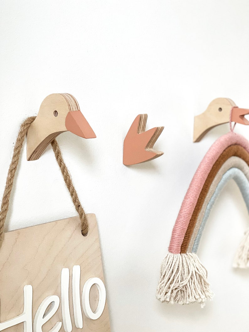 Op de afbeelding: Drie houten wandhaken in de vorm van ganzen en een hand, met een houten bord waarop "Hello" staat, dat aan een van de haken hangt. Een regenboogkleurige garenwandhanger hangt ook aan een van de haken.