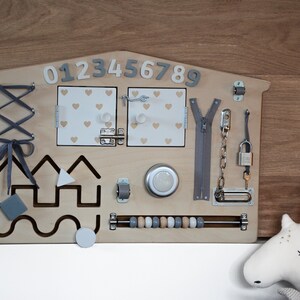 May include: Wooden activity board with various textures and activities for children, including a zipper, a lock and key, a latch, a lacing activity, a bead maze, and a shape sorter. The board is painted in a light wood color and has a house shape. The numbers 0123456789 are printed on the top of the board.