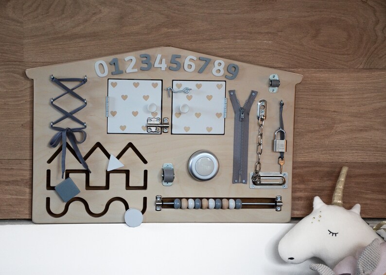 May include: Wooden activity board with various textures and activities for children, including a zipper, a lock and key, a latch, a lacing activity, a bead maze, and a shape sorter. The board is painted in a light wood color and has a house shape. The numbers 0123456789 are printed on the top of the board.