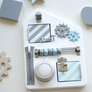 May include: A white wooden activity board with various textures and activities for children. It features a zipper, a latch, a rolling wheel, a knob, a set of gears, a sliding bead bar, and a small door with a latch.