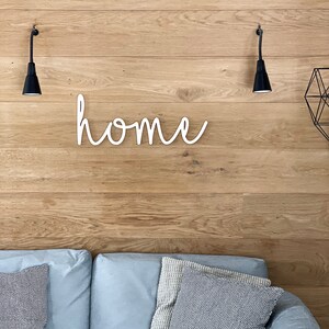 Może przedstawiać: Biały drewniany znak z napisem "home" napisanym kursywą, zamontowany na jasnej drewnianej ścianie z dwoma czarnymi kinkietami po obu stronach znaku. Szara sofa z poduszkami znajduje się na pierwszym planie.