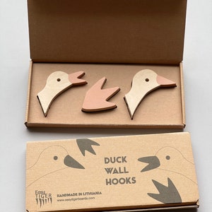 Op de afbeelding: Een set van drie houten eenden wandhaken in beige en roze. De haken zijn verpakt in een bruine kartonnen doos met de tekst "Duck Wall Hooks" en "Handmade in Lithuania" erop gedrukt.