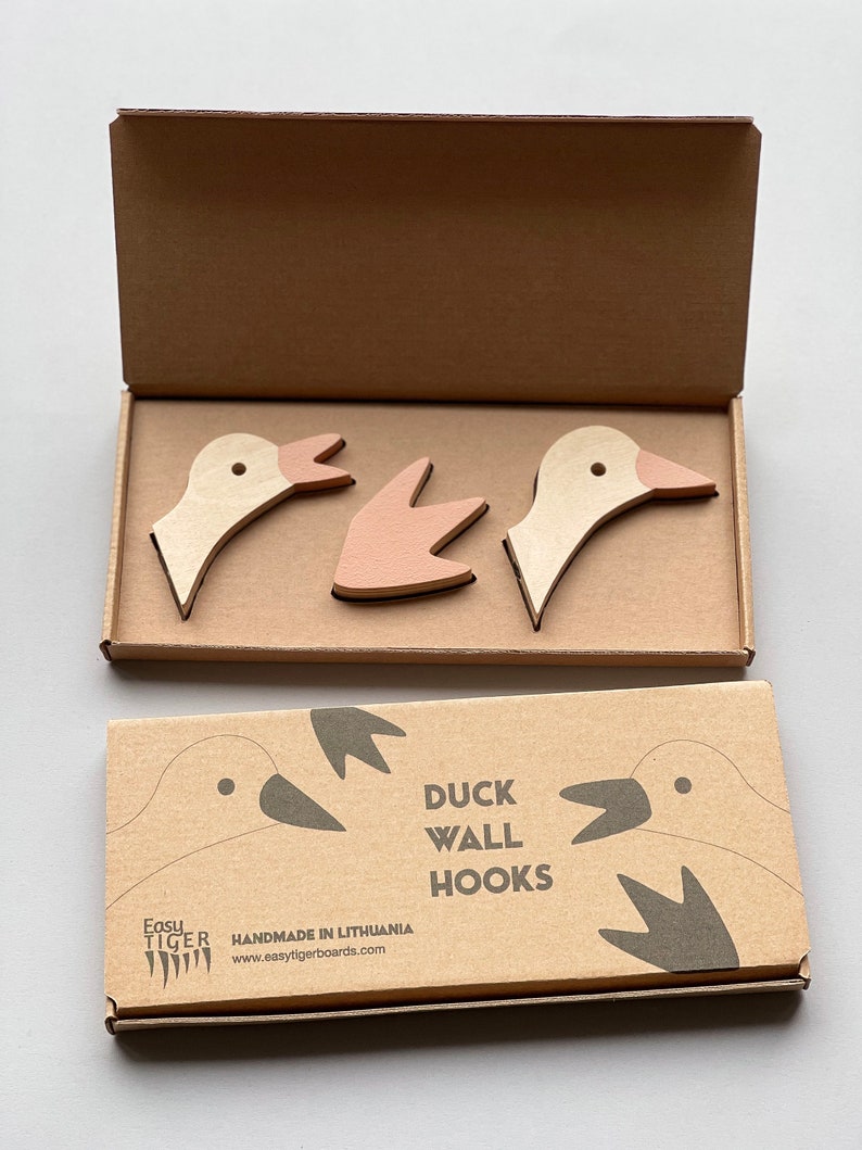 Op de afbeelding: Een set van drie houten eenden wandhaken in beige en roze. De haken zijn verpakt in een bruine kartonnen doos met de tekst "Duck Wall Hooks" en "Handmade in Lithuania" erop gedrukt.