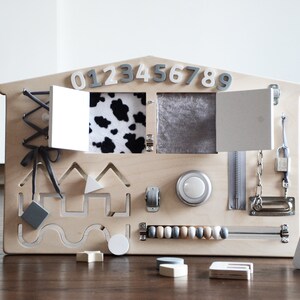 May include: Wooden activity board with various textures and activities for children, including a zipper, a latch, a lock, a chain, a bead maze, a cow print fabric flap, a gray fabric flap, a knob, a shape sorter, and a lacing activity. The board is painted in a light wood color and has the numbers 0123456789 and the letter A printed on it.