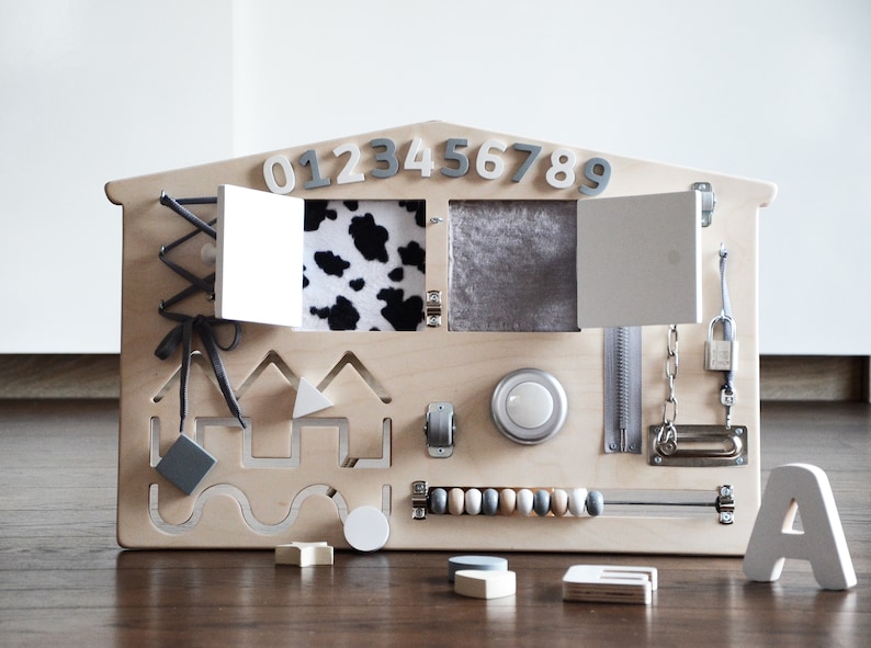May include: Wooden activity board with various textures and activities for children, including a zipper, a latch, a lock, a chain, a bead maze, a cow print fabric flap, a gray fabric flap, a knob, a shape sorter, and a lacing activity. The board is painted in a light wood color and has the numbers 0123456789 and the letter A printed on it.