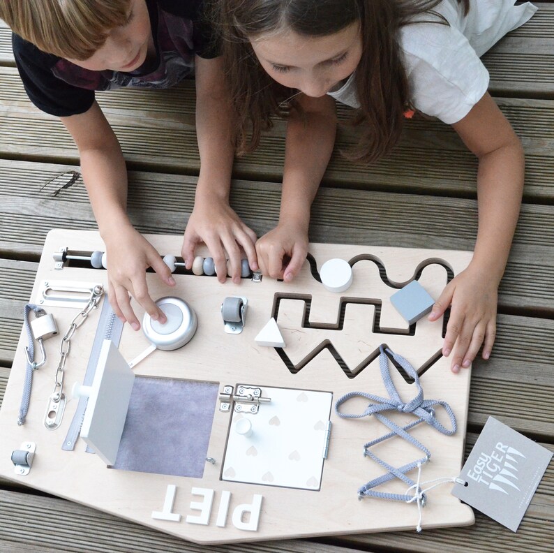 May include: A wooden activity board with various textures and activities for children. It features a latch, a zipper, a bell, a bead maze, a shape sorter, a knob, a sliding door, and a lacing activity. The board is labeled "PIET" and has a small tag with the text "EASY TIGER".